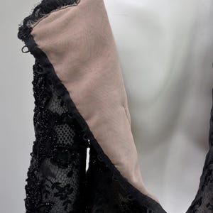 May include: Close-up of a black lace dress on a mannequin. The dress features intricate floral lace detailing, embellished with small, sparkling beads. The neckline and sleeves are visible, showcasing the delicate texture and design of the garment.