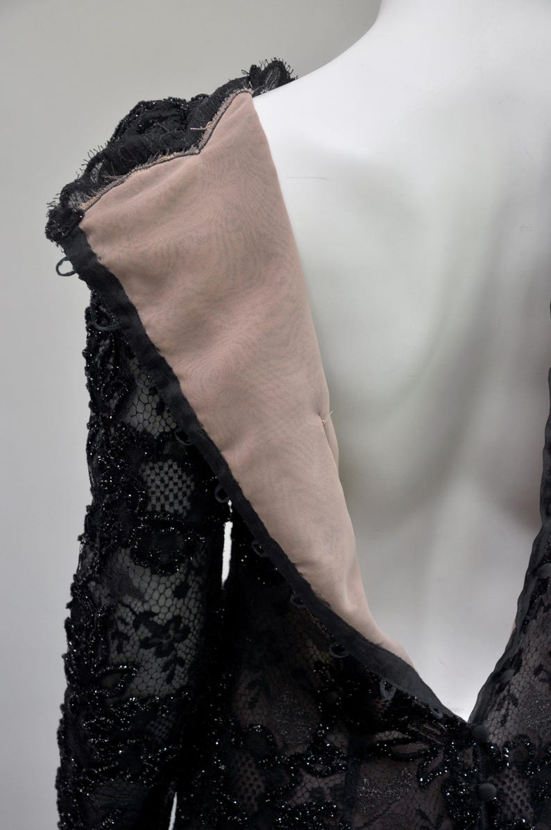May include: Close-up of a black lace dress on a mannequin. The dress features intricate floral lace detailing, embellished with small, sparkling beads. The neckline and sleeves are visible, showcasing the delicate texture and design of the garment.