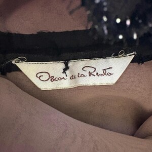 May include: Close-up of a garment label with the text "Oscar de la Renta" in brown script on a white tag. The tag is attached to a sheer, mauve fabric, with a black trim and a black, sequined detail at the top.