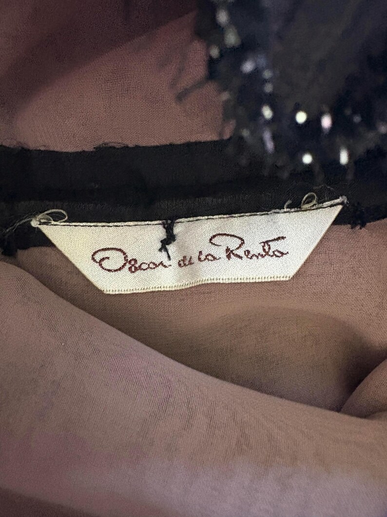 May include: Close-up of a garment label with the text "Oscar de la Renta" in brown script on a white tag. The tag is attached to a sheer, mauve fabric, with a black trim and a black, sequined detail at the top.