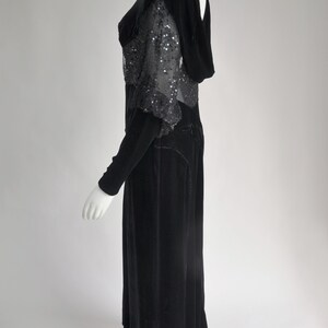 Puede incluir: Un vestido largo de terciopelo negro con mangas largas y capucha. El corpi&ntilde;o presenta una capa transparente con lentejuelas. El vestido es largo hasta el suelo y tiene una silueta ajustada, de estilo vintage.