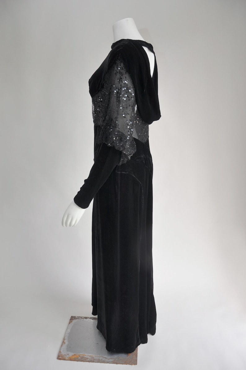 Puede incluir: Un vestido largo de terciopelo negro con mangas largas y capucha. El corpi&ntilde;o presenta una capa transparente con lentejuelas. El vestido es largo hasta el suelo y tiene una silueta ajustada, de estilo vintage.