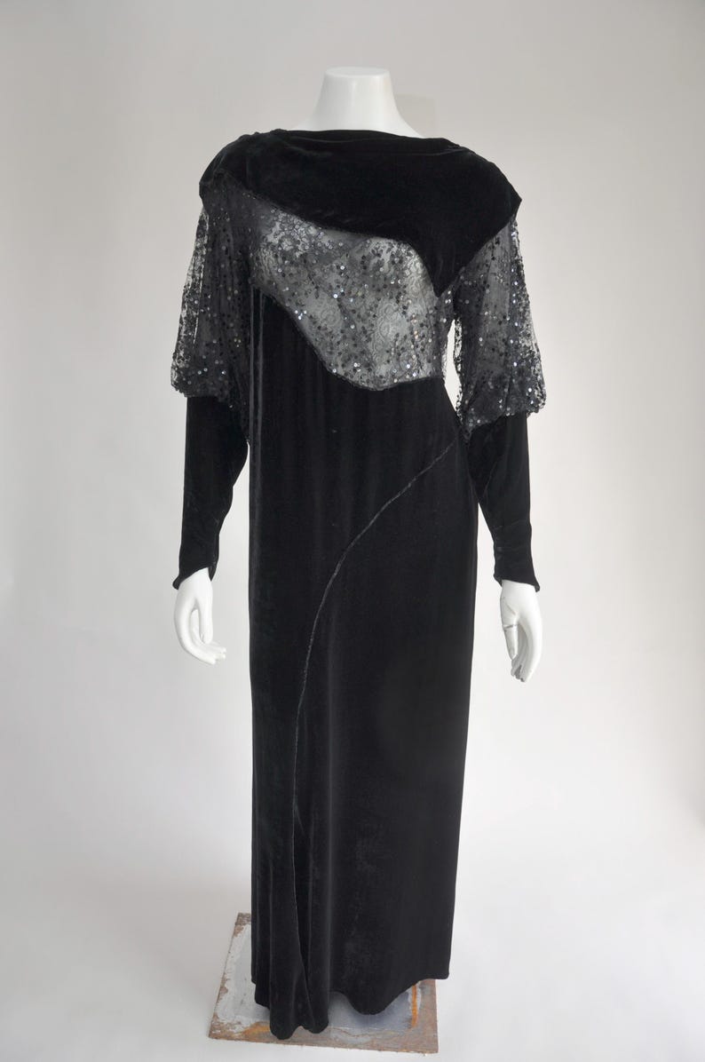 Puede incluir: Vestido de noche de terciopelo negro sobre un maniqu&iacute;. El vestido presenta mangas largas y una superposici&oacute;n de lentejuelas en el corpi&ntilde;o y las mangas superiores. Una costura diagonal recorre el frente.