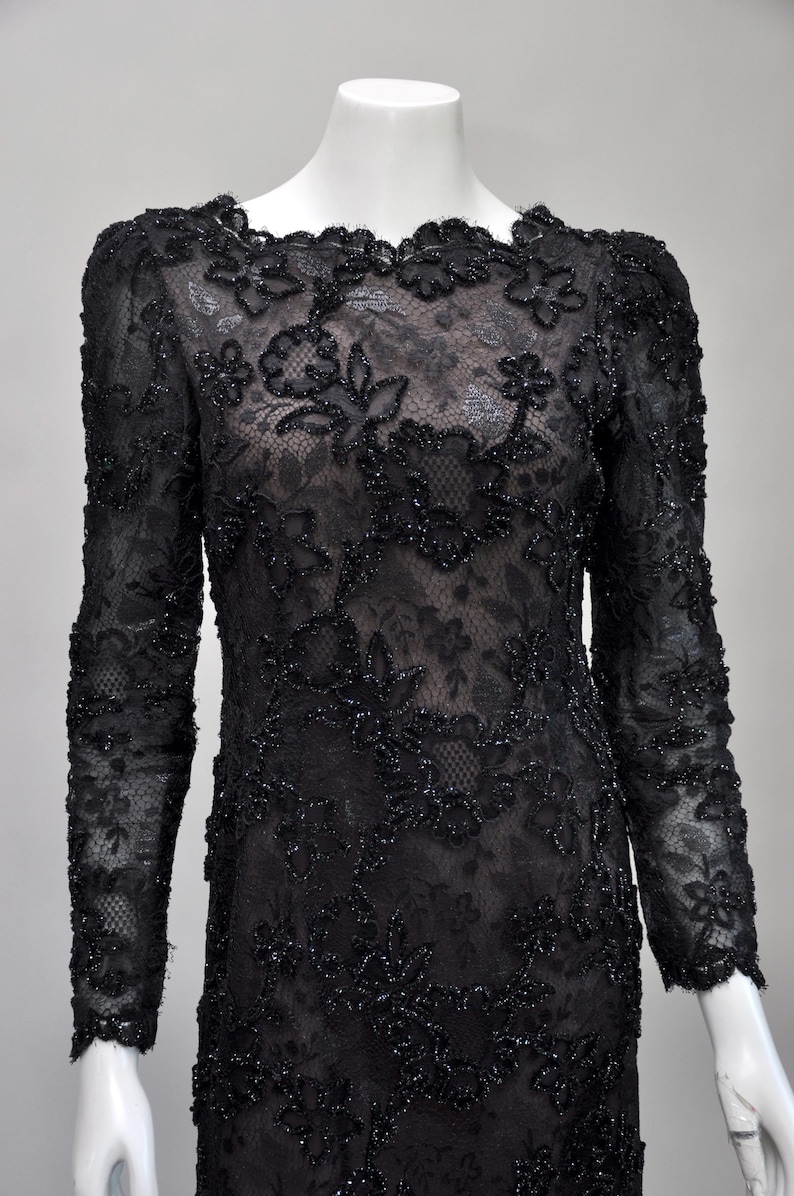 May include: Black lace dress with long sleeves and floral embroidery. The dress features a round neckline and is displayed on a mannequin. The intricate lace design is embellished with small, sparkling beads, creating a formal and elegant look.