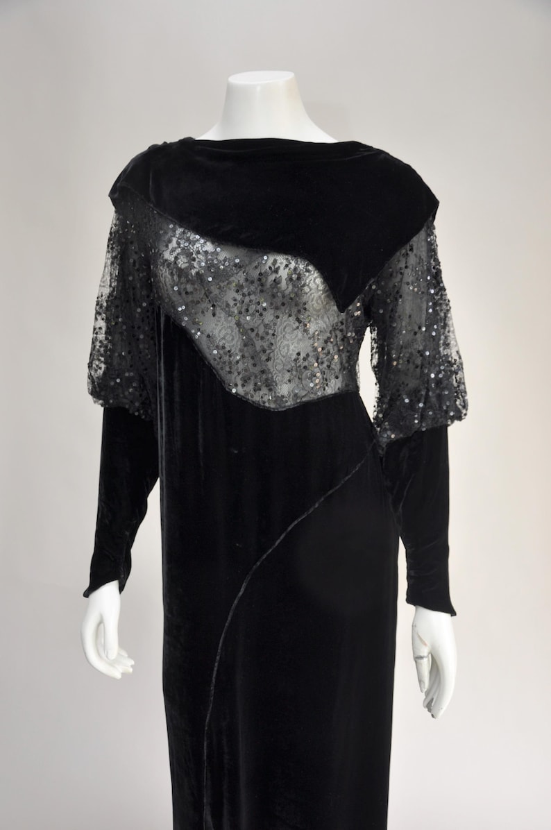 Puede incluir: Vestido de terciopelo negro con mangas largas y un dise&ntilde;o &uacute;nico. El vestido presenta un panel transparente con lentejuelas en el pecho y la parte superior de las mangas. El vestido se exhibe en un maniqu&iacute;.