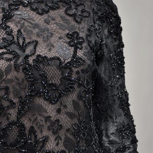 May include: Close-up of a black lace garment with floral patterns. The lace is embellished with sparkling accents, creating a textured and elegant appearance. The garment has long sleeves and a scalloped neckline, suggesting formal wear.