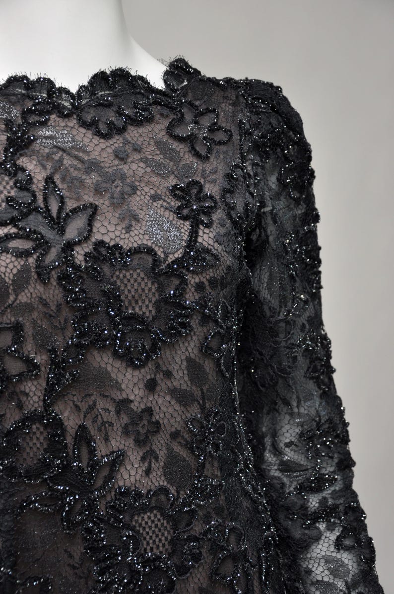 May include: Close-up of a black lace garment with floral patterns. The lace is embellished with sparkling accents, creating a textured and elegant appearance. The garment has long sleeves and a scalloped neckline, suggesting formal wear.