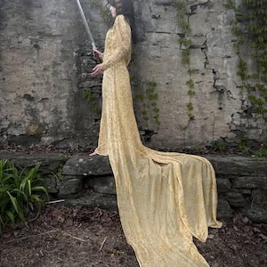 May include: A floor-length, long-sleeved gold dress with a textured pattern and a sweeping train. The garment features a fitted bodice and a high neckline, with the fabric draping elegantly over a stone ledge while holding a silver sword.