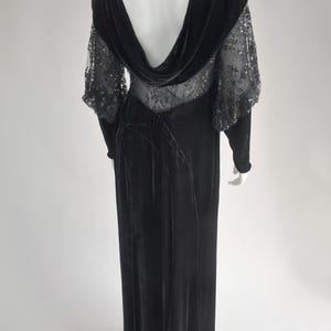 Puede incluir: Un vestido de noche de terciopelo negro con cuello alto y mangas transparentes con lentejuelas. El vestido presenta una falda larga y fluida y un dise&ntilde;o de espalda baja. El vestido se exhibe en un maniqu&iacute; sobre un fondo blanco.