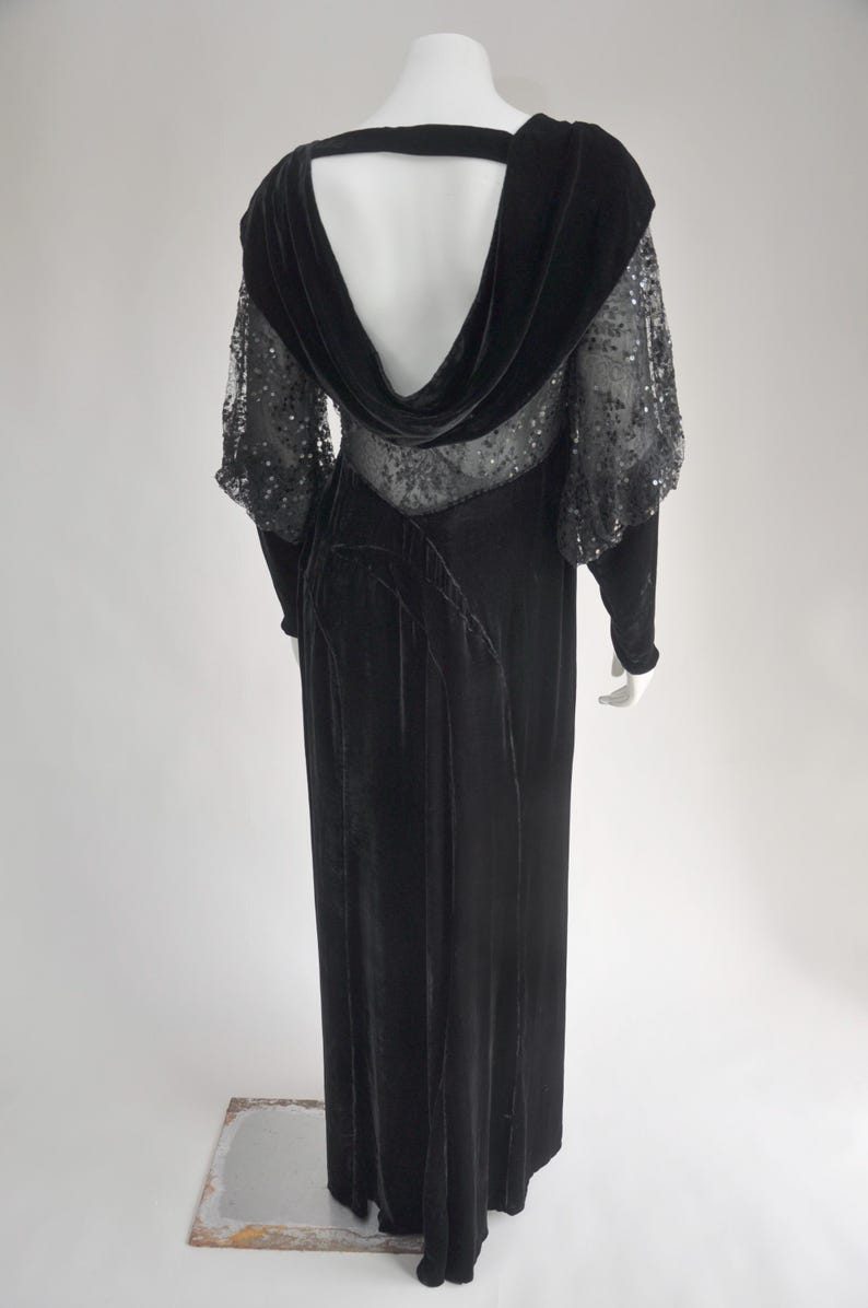 Puede incluir: Un vestido de noche de terciopelo negro con cuello alto y mangas transparentes con lentejuelas. El vestido presenta una falda larga y fluida y un dise&ntilde;o de espalda baja. El vestido se exhibe en un maniqu&iacute; sobre un fondo blanco.