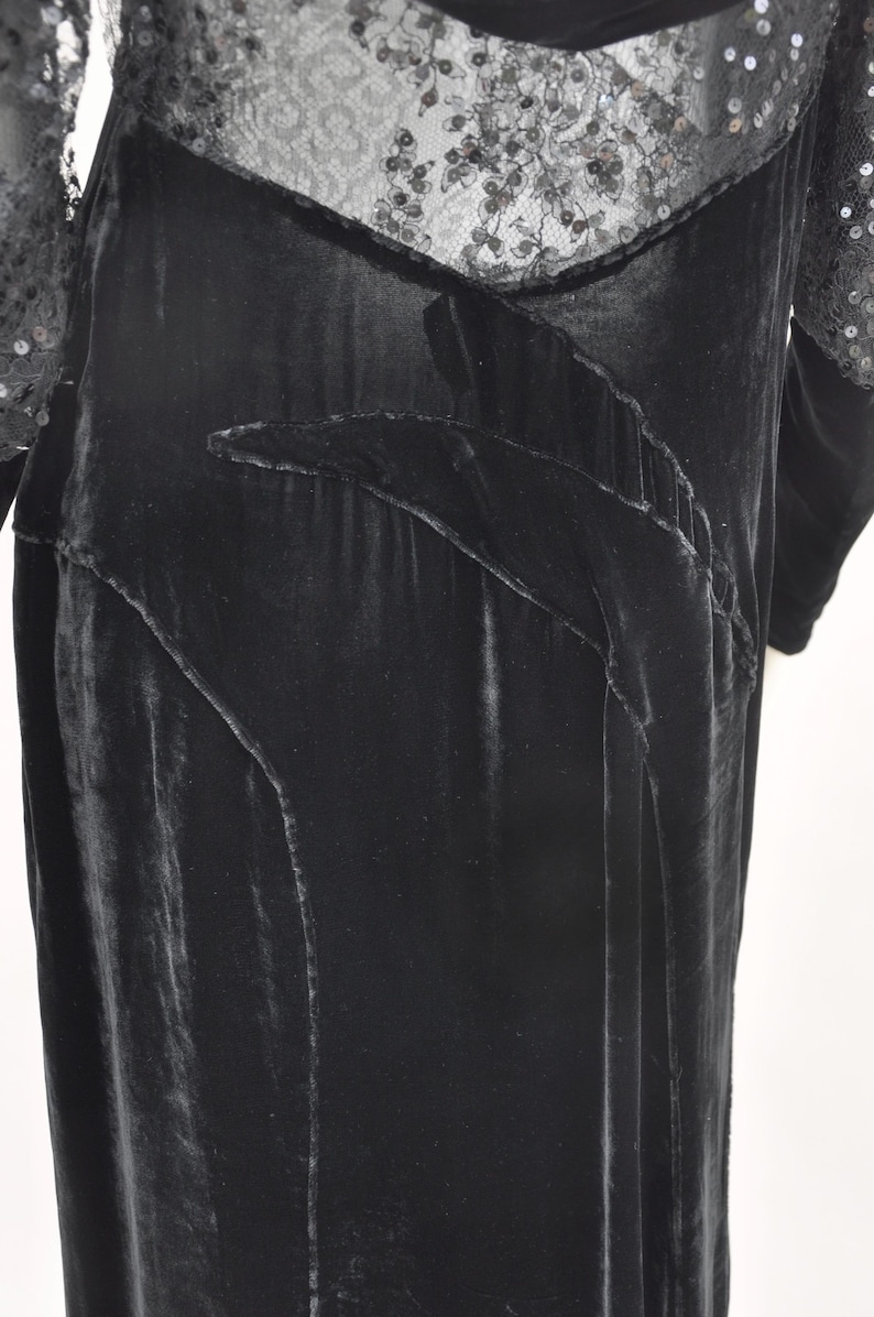 Puede incluir: Un vestido de terciopelo negro con escote y mangas de encaje con lentejuelas. El vestido presenta un dise&ntilde;o fluido con paneles decorativos y una textura suave y lujosa. Las mangas largas est&aacute;n adornadas con lentejuelas, a&ntilde;adiendo un toque de brillo.