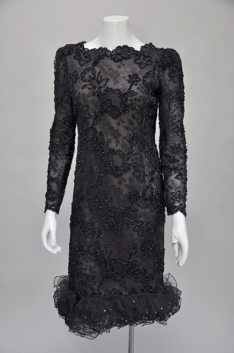 May include: Black lace dress with long sleeves and a ruffled hem. The dress features intricate floral embroidery and beadwork throughout. The neckline is scalloped, and the dress is displayed on a mannequin.