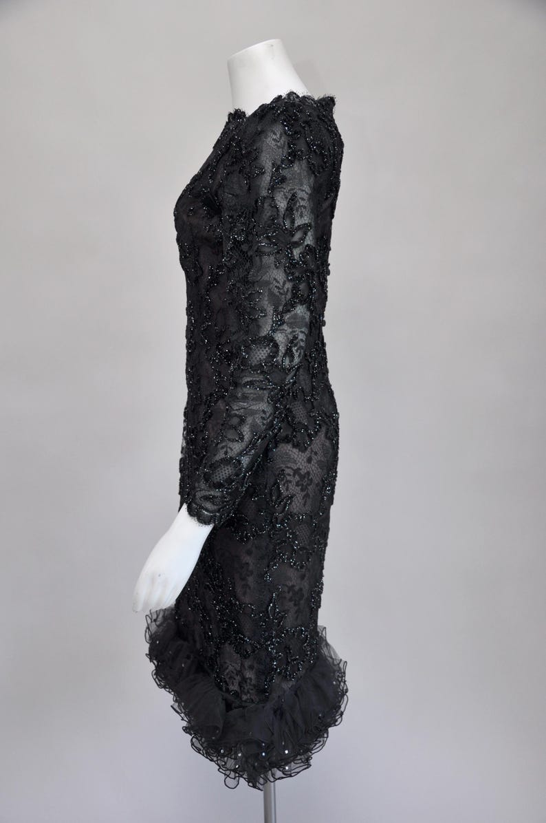 May include: A black, long-sleeved dress with intricate floral lace detailing. The dress features a fitted silhouette and a ruffled hemline. The fabric is embellished with small, sparkling accents, adding a touch of glamour to the formal attire.