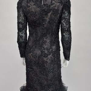 May include: A black, long-sleeved dress with intricate floral lace and bead embellishments. The dress features a ruffled hem and a row of buttons down the back. The dress is displayed on a mannequin.