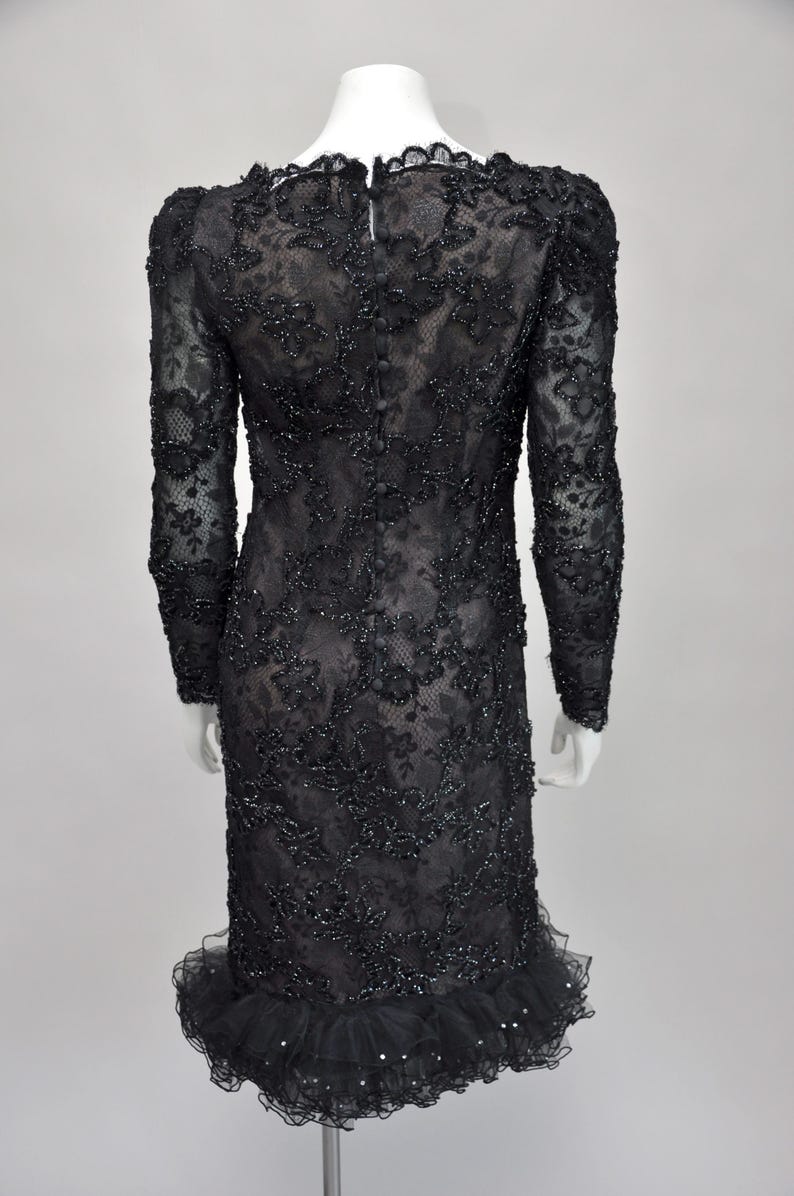 May include: A black, long-sleeved dress with intricate floral lace and bead embellishments. The dress features a ruffled hem and a row of buttons down the back. The dress is displayed on a mannequin.