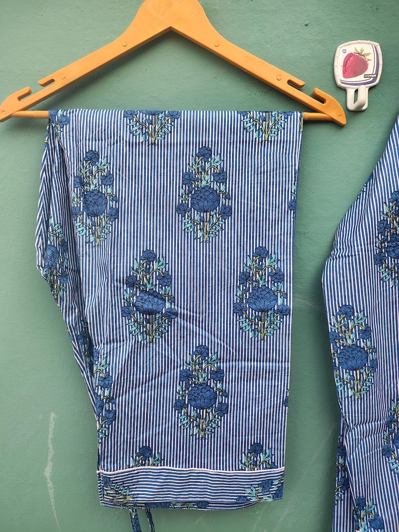 以下が含まれることがあります： 青と白のストライプのパジャマパンツ、花柄、木製のハンガーに吊るされています。パンツは青い花と葉の繰り返し模様が特徴です。裾には白い縁取りがあります。