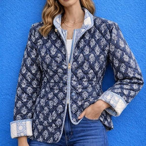 Può includere: Giacca trapuntata blu navy con motivo floreale bianco. La giacca ha una chiusura a zip, un collo alto e polsini con un design bianco e blu a contrasto. La modella indossa jeans blu.
