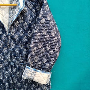 May include: A navy blue quilted jacket with a white floral pattern and vertical stripes. The jacket has a quilted lining, a notched collar, and button closures. The cuffs are folded up, revealing a white floral pattern. The jacket is hanging on a wooden hanger.