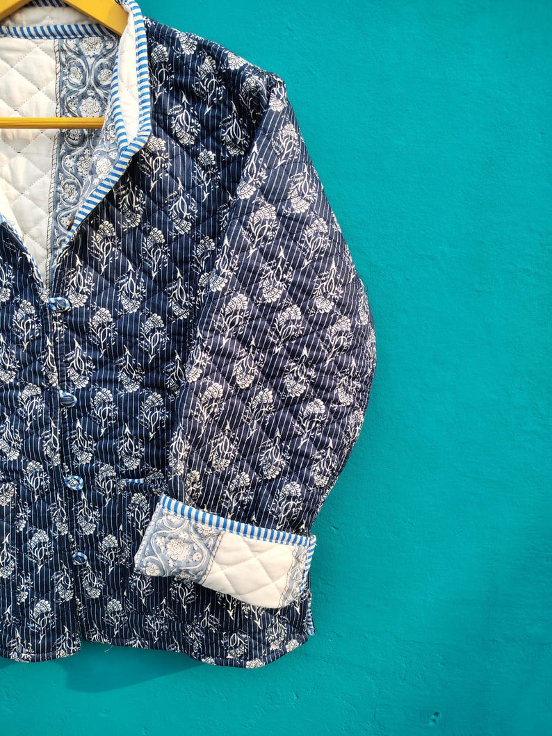 May include: A navy blue quilted jacket with a white floral pattern and vertical stripes. The jacket has a quilted lining, a notched collar, and button closures. The cuffs are folded up, revealing a white floral pattern. The jacket is hanging on a wooden hanger.