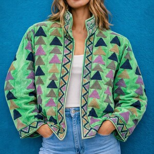 May include: A green jacket with embroidered triangle patterns in various colors, including purple, navy, and tan. The jacket has a high collar and a zigzag pattern along the edges. The model is wearing a white top and blue jeans.