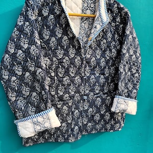 May include: A quilted jacket with a navy blue floral pattern and white accents. The jacket features a button-up front, a collar, and cuffs with a contrasting design. The jacket is hanging on a wooden hanger.