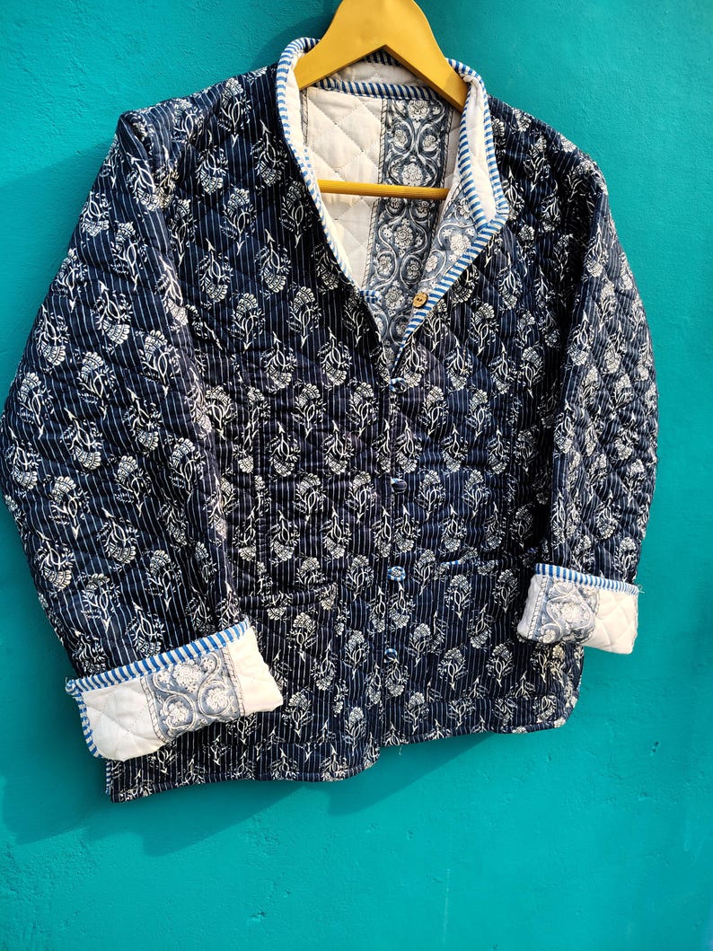 May include: A quilted jacket with a navy blue floral pattern and white accents. The jacket features a button-up front, a collar, and cuffs with a contrasting design. The jacket is hanging on a wooden hanger.