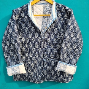 May include: A navy blue quilted jacket with a white floral pattern and white trim. The jacket has a button-up front, long sleeves, and a quilted design. The jacket is hanging on a yellow hanger.
