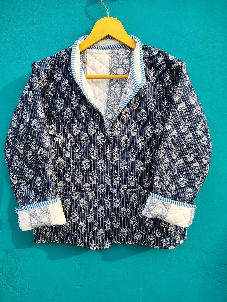 May include: A navy blue quilted jacket with a white floral pattern and white trim. The jacket has a button-up front, long sleeves, and a quilted design. The jacket is hanging on a yellow hanger.