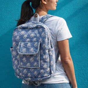 Puede incluir: Mochila azul con estampado floral, un bolsillo delantero y bolsillos laterales. La mochila tiene ribetes y correas a rayas blancas y azul marino. La bolsa está hecha de tela acolchada. El fondo es azul turquesa liso.