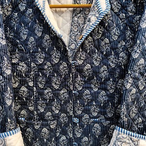 May include: A quilted jacket in navy blue with a white floral pattern and vertical stripes. The jacket has a mandarin collar, button closures, and white cuffs with a blue floral design. The interior is white.