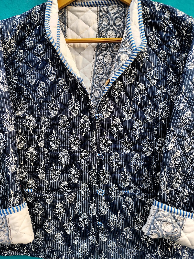 May include: A quilted jacket in navy blue with a white floral pattern and vertical stripes. The jacket has a mandarin collar, button closures, and white cuffs with a blue floral design. The interior is white.