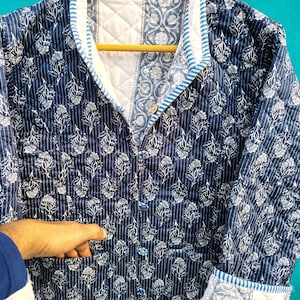 May include: A quilted jacket in navy blue and white floral print. The jacket features a button-up front, pockets, and a contrasting white collar and cuffs with blue stripes. The jacket is hanging on a wooden hanger.