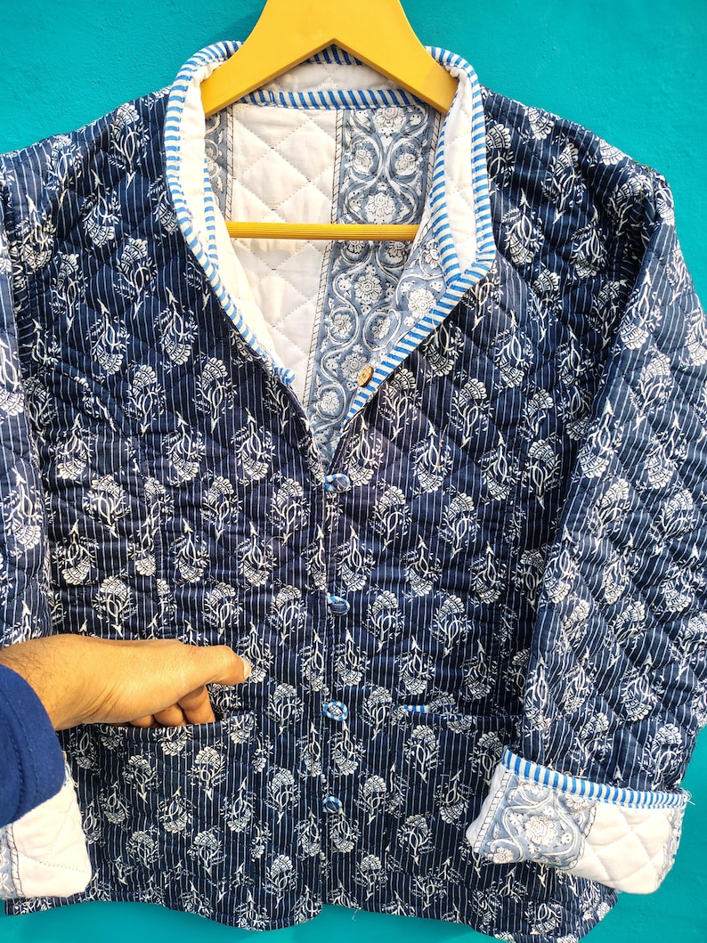 May include: A quilted jacket in navy blue and white floral print. The jacket features a button-up front, pockets, and a contrasting white collar and cuffs with blue stripes. The jacket is hanging on a wooden hanger.