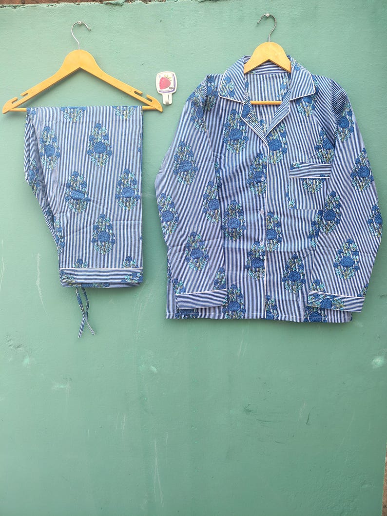 以下が含まれることがあります： 青と白のストライプのパジャマセット、花柄。 セットには、胸ポケット付きのボタンダウンシャツとパンツが含まれます。 パジャマは、緑色の背景に木製のハンガーに吊るされています。