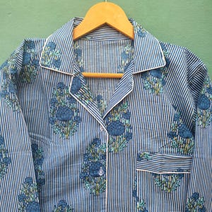 以下が含まれることがあります： 青と白のストライプのパジャマトップ、花柄。襟付き、胸ポケット、白いパイピングが特徴です。花柄は青と緑の色合いです。