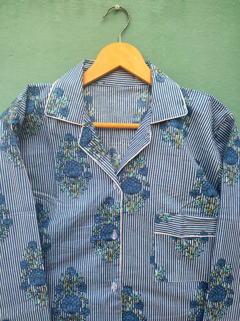 以下が含まれることがあります： 青と白のストライプのパジャマトップ、花柄。襟付き、胸ポケット、白いパイピングが特徴です。花柄は青と緑の色合いです。