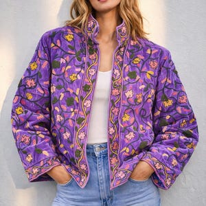 以下が含まれることがあります： 黄色、ピンク、緑の花柄刺繍が施された鮮やかな紫色のジャケット。 ジャケットはハイネックで、白いトップスと青いジーンズと合わせています。 ジャケットのデザインは、繰り返しの花柄が特徴です。