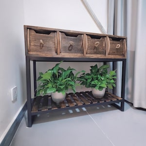 May include: A wooden storage unit with four drawers and a lower shelf, supported by a black metal frame. The drawers have rope handles. Two potted green plants sit on the lower shelf, adding a touch of nature.