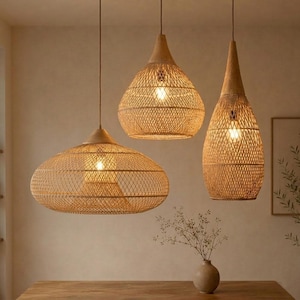 Op de afbeelding: Drie hanglampen met geweven rotan kappen hangen aan het plafond. De kappen hebben verschillende vormen: een brede ovaal, een afgeronde druppel en een lange, slanke druppel. Elke lamp heeft een zichtbare gloeilamp en een natuurlijke, lichtbruine kleur.