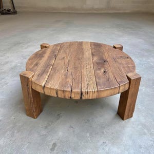 May include: A circular coffee table crafted from light brown wood, showcasing a rustic aesthetic. The tabletop is made from wooden planks, displaying natural grain and cracks. It stands on four square legs, offering a low design. Ideal for a living room or lounge space.