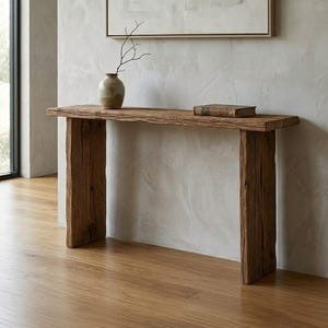 Puede incluir: Una consola rústica de madera con un acabado natural y envejecido. La mesa presenta una gruesa tapa rectangular sostenida por dos robustas patas verticales de madera. Un pequeño jarrón con una rama y un libro descansan sobre la superficie de la mesa. La mesa está contra una pared neutra.