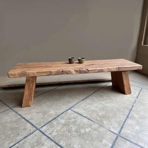 Puede incluir: Una mesa de centro rectangular de madera rústica con un aspecto natural sin terminar. La mesa tiene una gruesa tapa de borde vivo y patas anguladas. Dos cuencos pequeños y oscuros están colocados sobre la superficie. La mesa es de madera marrón claro.