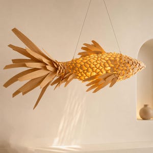 Può includere: Una lampada a sospensione a forma di pesce, in legno chiaro. Il corpo è fatto di pezzi di legno stratificati, creando un effetto a scaglie. Le pinne e la coda sono composte da lunghe doghe di legno appuntite. La luce brilla attraverso il corpo.