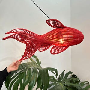 以下が含まれることがあります： 織り素材で作られた、赤い魚の形をしたペンダントライト。照明器具は黒いコードから吊り下げられており、内部に電球があります。魚にはひれと尾があり、白い壁と緑の植物の前に吊り下げられています。