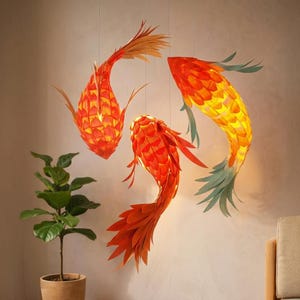 May include: Three hanging fish-shaped lamps in shades of orange and yellow. The lamps feature intricate fin and tail details, with a layered design. They are suspended from the ceiling, providing decorative lighting. A potted plant is in the lower left corner.