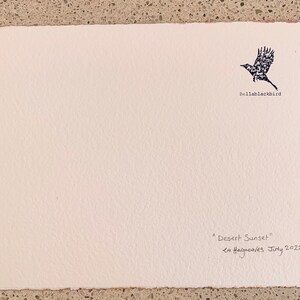 May include: A white card with a black silhouette of a bird flying, the text "Bellablackbird" below the bird, and the text "Desert Sunset" and "La Haigreares July 2022" handwritten in black ink at the bottom.