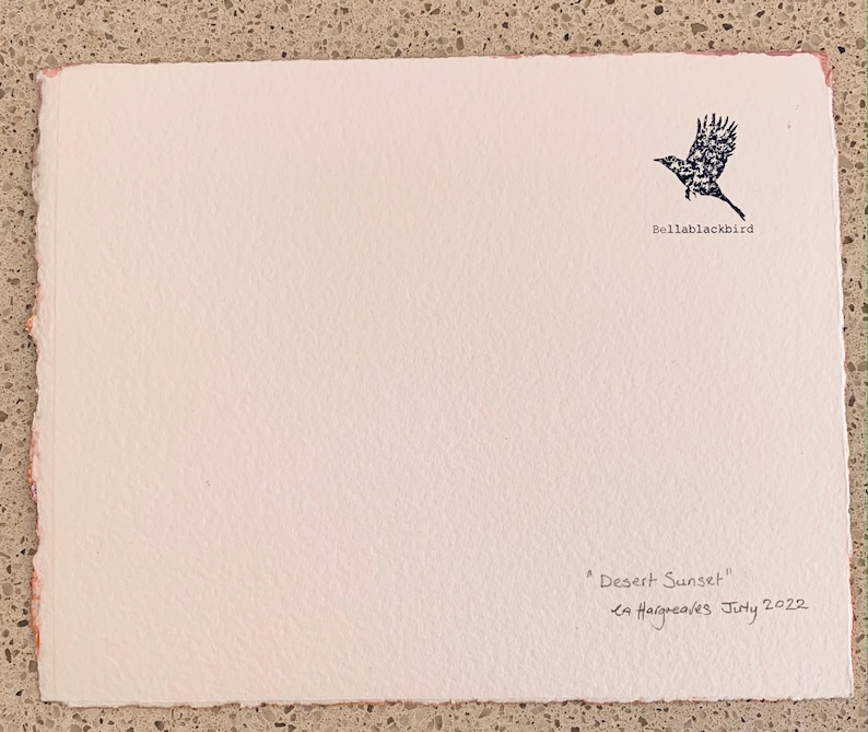 May include: A white card with a black silhouette of a bird flying, the text "Bellablackbird" below the bird, and the text "Desert Sunset" and "La Haigreares July 2022" handwritten in black ink at the bottom.