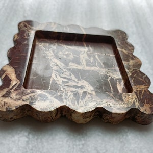 May include: A square-shaped tray with a wavy edge, made of brown and beige marble. The tray has a raised inner rim and a smooth, polished surface.