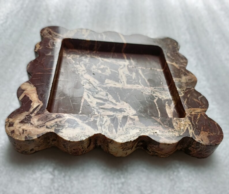 May include: A square-shaped tray with a wavy edge, made of brown and beige marble. The tray has a raised inner rim and a smooth, polished surface.