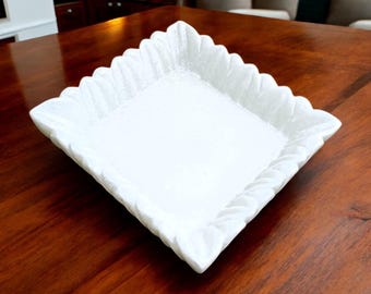 May include: White square serving tray with a decorative carved edge. The tray is made of marble and has a smooth, polished surface.